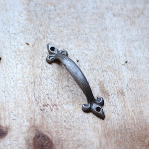 May include: A silver-toned metal drawer pull with a decorative, ornate design. The pull has a curved shape and a pointed end.