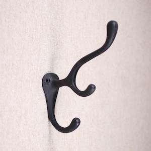 May include: A black cast iron coat hook with three curved arms. The hook is mounted on a wall and has a matte finish. The design is simple and functional, suitable for entryways or bedrooms.