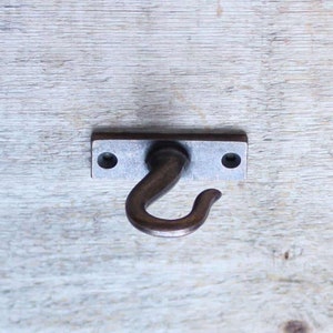 Peut inclure: Un crochet en métal argenté avec un design incurvé est fixé à une plaque rectangulaire en métal. Le crochet est monté sur une surface en bois clair.