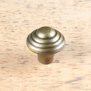 Peut inclure: Un bouton de meuble de couleur bronze avec un design circulaire en couches. Le bouton a un dessus arrondi et une base courte et cylindrique. La quincaillerie est probablement en métal et est conçue pour être utilisée sur des tiroirs ou des portes d'armoires.