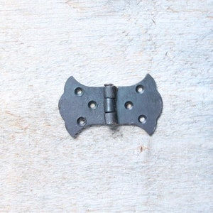 May include: Black metal hinge with a decorative, ornate design. The hinge has four holes on each side for attaching to a door or cabinet.