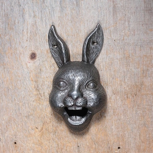 May include: A black cast iron rabbit bottle opener with a smiling face and a bottle opener in its mouth.
