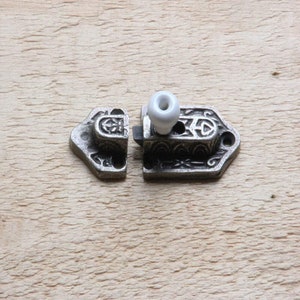 Peut inclure: Un loquet en métal argenté avec un motif orné et un bouton en céramique blanche. Le loquet est en deux parties, le bouton étant fixé sur le côté droit.
