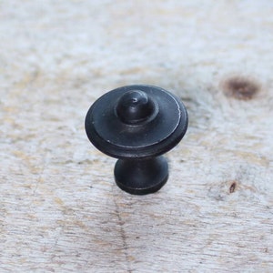 Op de afbeelding: Zwarte metalen kastknop met een ronde bovenkant en een licht verhoogd midden. De knop heeft een eenvoudig, klassiek ontwerp.