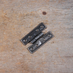 May include: A black metal hinge with a rectangular shape and rounded corners. The hinge has multiple screw holes and a central joint. The hinge is open, showing the connection between the two parts. The background is a light brown wooden surface.