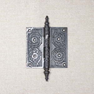 Könnte beinhalten: Ein dekoratives Scharnier im antiken Stil aus dunklem Metall. Das Scharnier weist verzierte, wirbelnde Muster und einen zentralen Stift auf. Die rechteckigen Platten haben kreisförmige Designs und sind durch einen detaillierten Mittelteil verbunden.