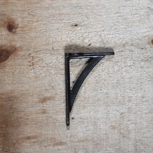 May include: Black metal shelf bracket with a curved design. The bracket is mounted on a light brown wooden surface.