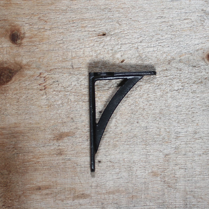 May include: Black metal shelf bracket with a curved design. The bracket is mounted on a light brown wooden surface.