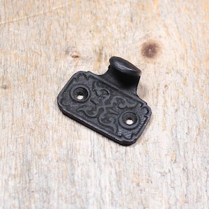 May include: A black metal sash lift featuring a rectangular base with an ornate, raised scrollwork pattern and two countersunk screw holes. The hardware includes a curved, protruding handle grip extending from the top edge of the plate.