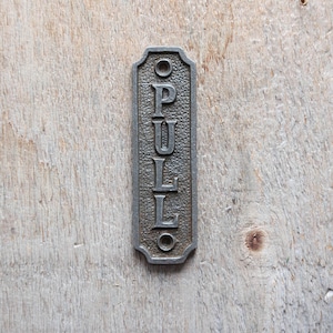 Op de afbeelding: Een zilveren metalen bord met het woord "PULL" erop gegraveerd. Het bord is bevestigd aan een houten oppervlak.