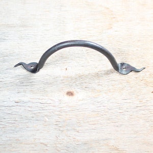 Könnte beinhalten: Ein schwarzer Metalltürgriff mit gebogenem Design und spitzen Enden. Der Griff ist auf einer hellholzfarbenen Oberfläche montiert.
