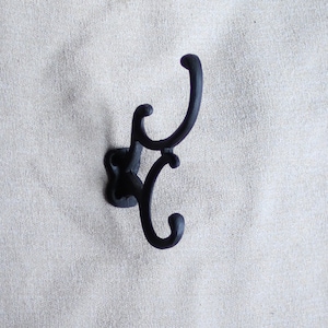 Può includere: Un gancio da parete in ghisa nera con un design decorativo. Il gancio ha tre punte.