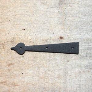 May include: A black metal hinge with a decorative heart-shaped end and three holes for mounting. The hinge is shown against a light wood grain background. The hinge measures approximately 25 cm in length.