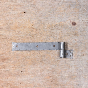 May include: A silver metal hinge with a decorative hammered finish. The hinge is bent at a 90-degree angle and has four holes for attaching to a door or gate.
