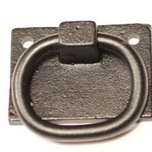 Op de afbeelding: Een zwarte gietijzeren ringgreep met een vierkante montageplaat. De ring is ovaal en bevestigd aan de plaat met een vierkante basis. Twee gaten zijn zichtbaar op de plaat voor montage. De hardware heeft een getextureerde afwerking.