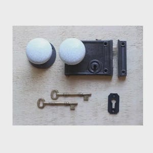 May include: A black antique door latch set with two white crackle-glazed ceramic knobs, two brass keys, and a black keyhole cover. The set is designed for a vintage or rustic aesthetic.