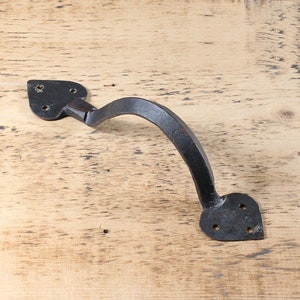 May include: A black, wrought iron handle with a curved grip. The handle has heart-shaped mounting plates with pre-drilled holes for screws. The metal has a textured, rustic appearance, suggesting a handmade or antique quality.