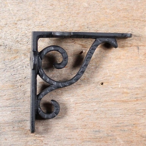 Peut inclure: Équerre d'étagère en fer forgé noir avec un motif décoratif en volutes. Le support a un dessus plat et une forme en angle droit, adapté pour soutenir des étagères. Le métal a un aspect texturé et martelé.