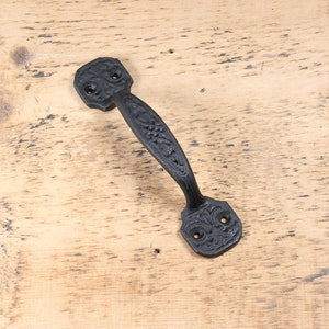 May include: Black cast iron door handle with a floral design. The handle has a curved shape and two holes for mounting.