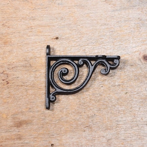 May include: A black, cast iron shelf bracket with an ornate, scrolled design. The bracket is L-shaped, with a flat top and a vertical mounting plate. The decorative scrolls add a vintage, antique aesthetic. The bracket is shown against a light wood background.