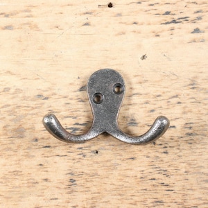 Op de afbeelding: Een dubbele haak van verouderd metaal. De haak heeft een afgeronde bovenkant met twee schroefgaten en twee gebogen haken. Het metaal heeft een gevlekte, grijze afwerking en is geplaatst tegen een lichte houten achtergrond.