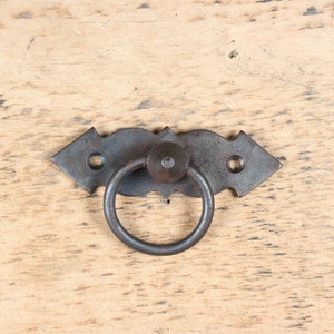 May include: Black metal ring pull handle with a decorative design. The handle is attached to a metal plate with two screws.