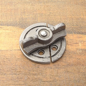 Può includere: Un chiavistello circolare in ghisa con una maniglia a leva e due fori per viti. Il chiavistello è di colore grigio scuro e si trova su una superficie di legno. La maniglia a leva è progettata per una presa e un funzionamento facili.