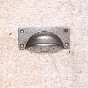May include: A cast iron drawer pull with a rounded, recessed handle. The pull is a dark gray color and has a rectangular shape.