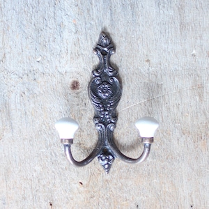 May include: A double hook with a decorative, ornate design in a dark bronze finish. The hooks have white ceramic knobs.