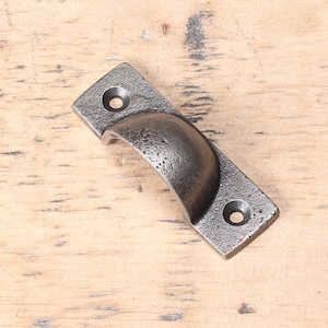 May include: A cast iron cabinet pull with a textured, dark gray finish. The rectangular base has two holes for mounting, and the curved handle provides a comfortable grip. This rustic hardware is suitable for drawers and cabinets.
