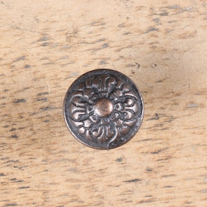May include: A round, ornate, copper-coloured knob with a floral design. The knob is set against a light brown wooden surface.