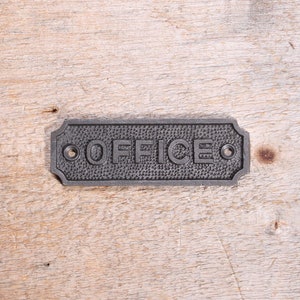Peut inclure: Une plaque en métal noir avec le mot "OFFICE" en relief. La plaque a deux petits trous de chaque côté.