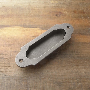 Peut inclure: Une poignée de porte en métal gris avec une ouverture rectangulaire. La poignée a une forme légèrement arrondie et est conçue pour être montée à fleur.