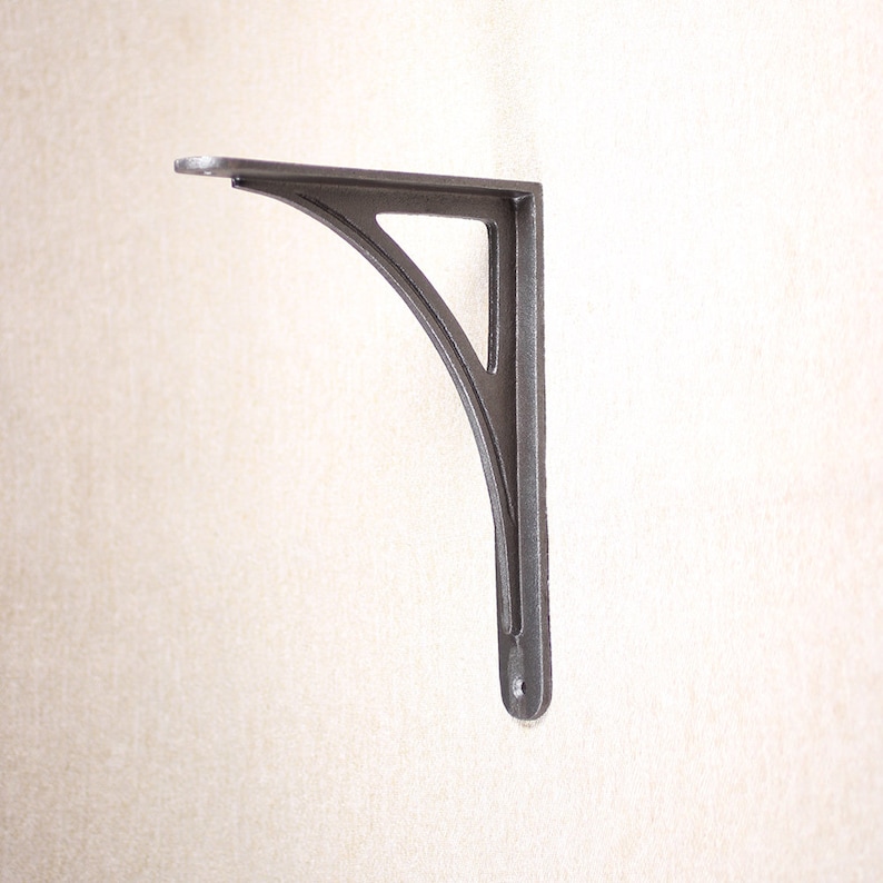 May include: A dark gray, cast iron shelf bracket. The bracket has a curved design with a triangular cutout and a flat top. It is designed to be mounted on a wall and support a shelf. The bracket is made of metal and has a rustic, industrial look.