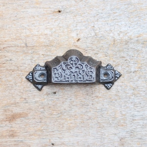 Op de afbeelding: Een zwarte gietijzeren ladegreep met een sierlijk ontwerp. De greep heeft een gebogen bovenkant en twee decoratieve uiteinden.
