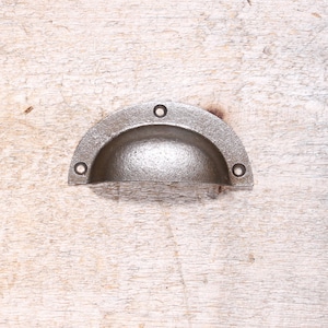 May include: A cast iron cup pull with a semi-circular design. The handle has a textured, matte finish and three holes for mounting. The hardware is a muted gray color and is designed for use on drawers or cabinets.