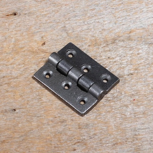 May include: A dark grey metal hinge, rectangular with rounded corners. It features screw holes and a central barrel for articulation. The hinge is displayed on a wooden surface.