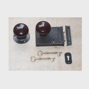 May include: A vintage door hardware set comprising two dark brown doorknobs, a black lock mechanism, a strike plate, and two brass keys. The set includes a keyhole cover, all arranged on a light wood surface.