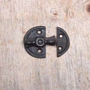 May include: A black cast iron cabinet latch with a half-moon design. The latch has a rotating handle and is mounted on a light-coloured wooden surface. The hardware is designed for securing cabinet doors or small boxes.