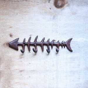 May include: A brown metal fish skeleton shaped wall hook with five hooks. The hook is mounted on a light wood surface.