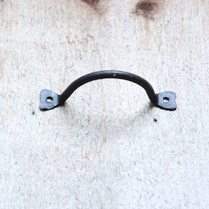 May include: Black metal handle with a curved shape and rounded ends. The handle is attached to a wooden surface.