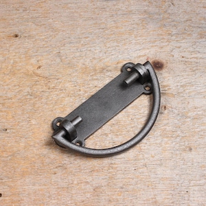 May include: A black metal door handle with a rectangular plate and a rounded pull handle. The handle has a rustic, industrial look.