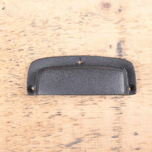 May include: A black, cast iron drawer pull with a textured surface. The pull has a curved shape with two mounting holes on each end and a recessed center for gripping. Hardware for furniture.