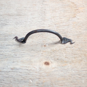 May include: A dark metal handle with a curved bar and decorative, bird-like ends. This handle is designed for furniture or cabinetry, offering a vintage aesthetic. The metal has a slightly textured appearance, suggesting a hand-forged quality.