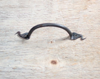 May include: A rustic, dark metal handle with a curved bar and decorative, bird-like ends. The handle is designed for furniture or cabinetry, offering a vintage aesthetic. The metal has a slightly textured appearance, suggesting a hand-forged quality.