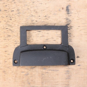 May include: Black metal drawer pull with a rectangular cutout for a label. The pull has a rounded bottom and two small holes on each side.