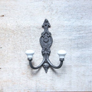 Könnte beinhalten: Ein dekorativer, verzierter Doppel-Haken für die Wand. Die Metallhalterung ist dunkelgrau mit einem floralen Design und zwei weißen Keramikknöpfen. Der Haken ist auf einer hellen, strukturierten Holzoberfläche montiert.