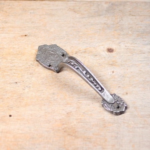 May include: A silver cast iron door handle with an ornate design. The handle has a floral pattern and a decorative shield shape at the top.