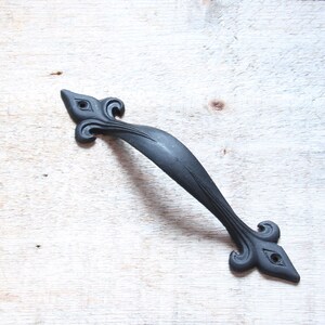 May include: A black, ornate door pull with a fleur-de-lis design. The handle is made of metal and has a matte finish. The decorative hardware is set against a light wood background.