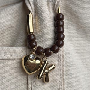May include: A gold-coloured bag charm featuring a heart pendant, a letter "K" charm, and a brown beaded accent. The heart charm has a small, clear gem. The charm is attached to a beige canvas bag.